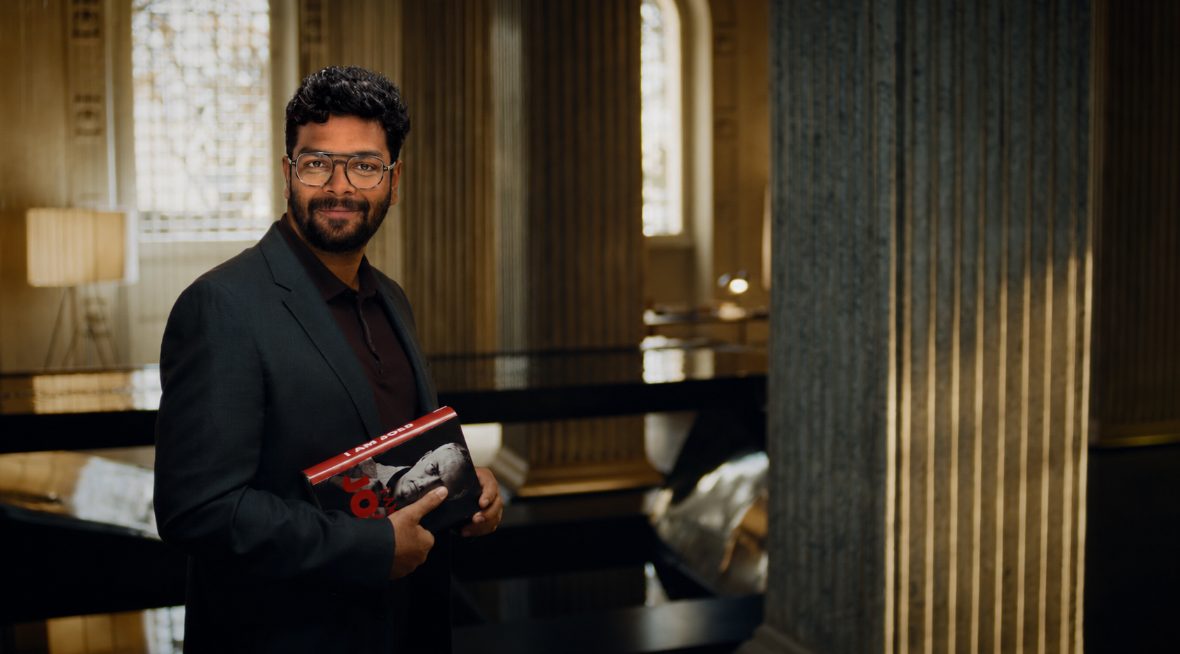 Suhail Shaikh standing in a wood-paneled hall, holding a book